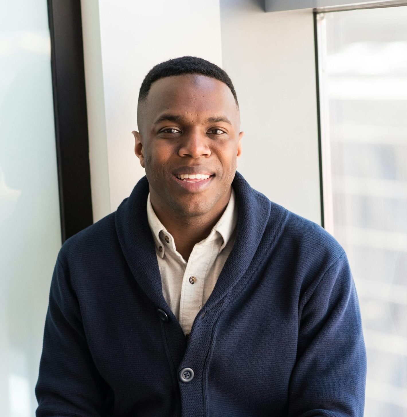 A professional black man smiling while seated by a window, embodying modern business vibes.